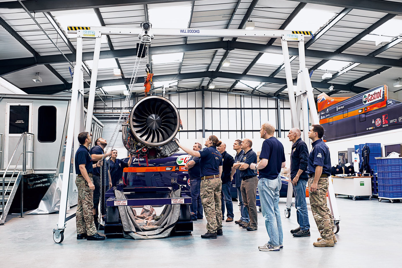 Group of people in a hangar with a large aircraft engine hanging from gantry lifting device.